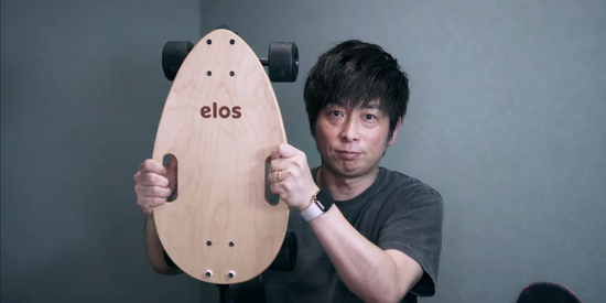 【ミニ クルーザー おすすめ】男性がElosを持ち上げ、Elosの楽しさを紹介しています。Elosは多くの方におすすめの ミニクルーザーで、カーバースケートボードやロングスケートボードの代替品です。Elosはプレミアムな スケートボードパーツを使用しています。
